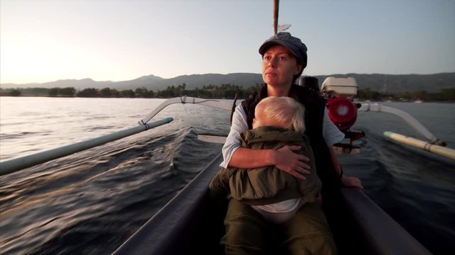 Caucasian mother is holding two-years old son while sailing in traditional indonesian canoe, motor jukung, surching for dolphins in the morning. Bali, Indonesia