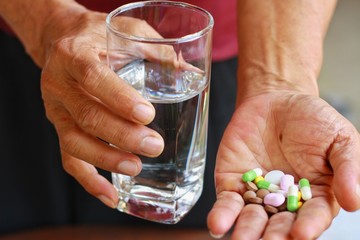 Elderly Asian man holding many pills(tablet,capsule ) and glass of water in hands.Taking a lot of medicine, supplements or antibiotic antidepressant painkiller medication.Hope for cure,close up view.