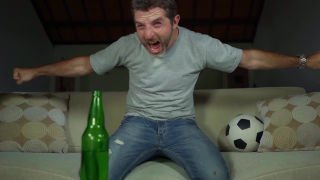 young attractive excited soccer fan man nervous at home couch with beer watching football match celebrating goal crazy happy dancing and gesturing on living room sofa feeling the emotion of victory