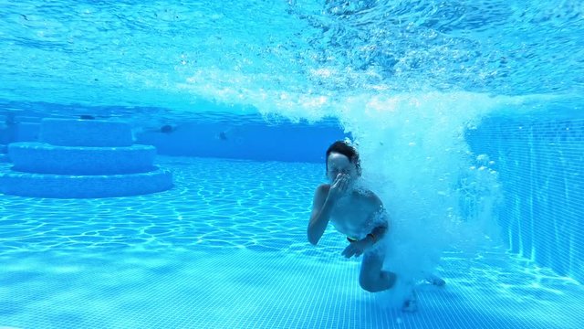 Happy boy jumping into clear water in the swimming pool outdoors. Young guy jumps underwater and swims with closed nose in blue background.