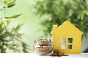 Model of house and jar with coins on table against blurred green background. Space for text