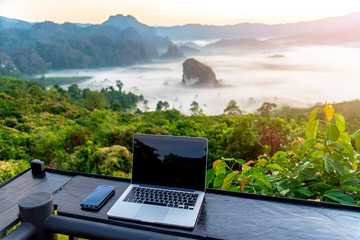A laptop, mobile  on wooden table with sunrise and mountain fog background in morning. A start of...