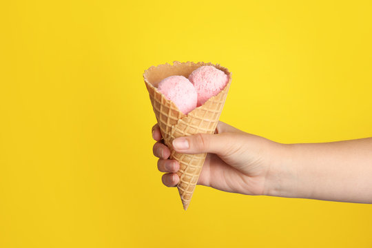 Woman holding delicious ice cream in wafer cone on yellow background, closeup