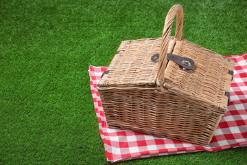 Checkered tablecloth and closed wicker picnic basket on green grass, space for text