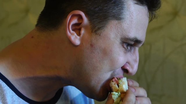 Cute young hungry caucasian man with an appetite is eating a tasty and juicy sandwich, carefully chewing up a close-up, slow motion
