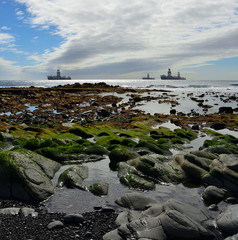 Fototapeta na wymiar Rocky beach with puddles at low tide in foreground, cloudy sky and oil rigs in background 