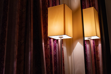 Modern rectangle lampshade and reflection in mirror on wall with red ornate curtains. Golden light...