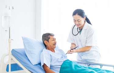 Obraz premium Asian woman professional doctor with clipboard visiting, talking, and diagnosing the old man patient lying in patient’s bed at hospital ward