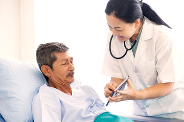 Asian woman professional doctor with clipboard visiting, talking, and diagnosing the old man patient lying in patient’s bed at hospital ward