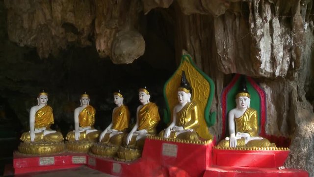 Handheld, interior, medium wide shot of a stalactite ceiling of a cave, then tilting down to reveal six Buddha statues.