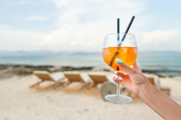 Woman holding Delicious classic iced aperol spritz cocktail with ice cubes on a hot tropical beach in summer sunshine