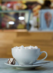 Mug of coffee with marshmallows on porcelain plate on blurred colorful interior bokeh background.