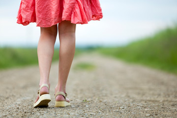 Back view of young woman in red dress legs walking by ground road on summer day on blurred sunny background.