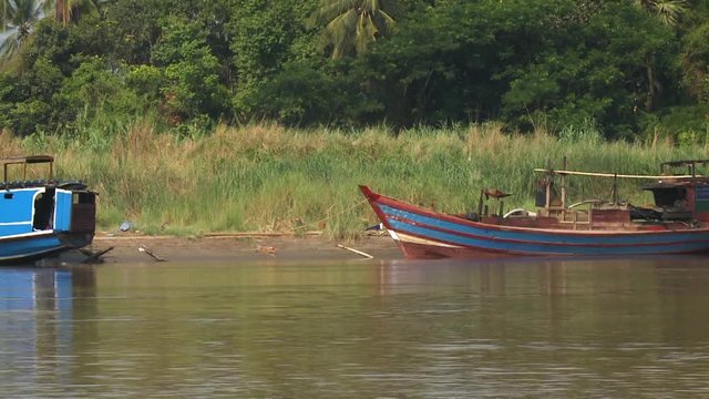 A daylight closeup shot from inside a moving passenger boat traveling in the murky river water passing by some motorboats and pump boats parked by the riverside.