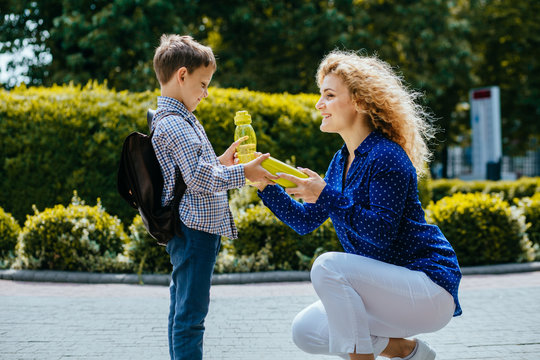 Blond curly caucasian mother giving school lunch and bottle with wather to her little son outdoors in sunny morning. Back to school concept.