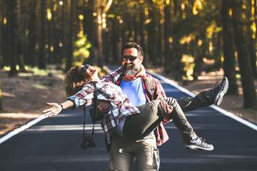 Fototapeta na wymiar Smiling romantic couple walking in the middle of the street having fun outdoors in a mountain forest. Trendy hipster tourist enjoying adventurous vacation together. Travel adventure and love concept