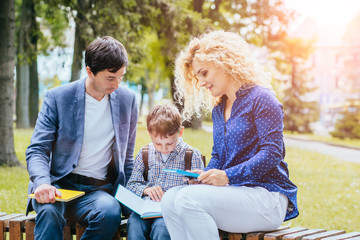 Fototapeta premium Curly blond mother and father are sittting on bench and teaching pupil kid in park with book outdoor. Happy family together concept.