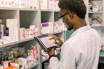 African American male pharmacist making notes on digital tablet in pharmacy, drug quality control