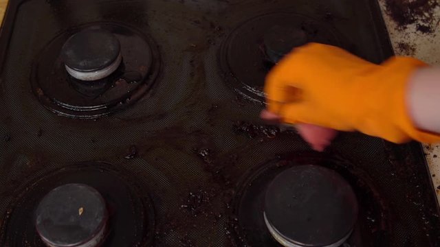 Close-up of hands in orange gloves cleaning dirty black gas stove.