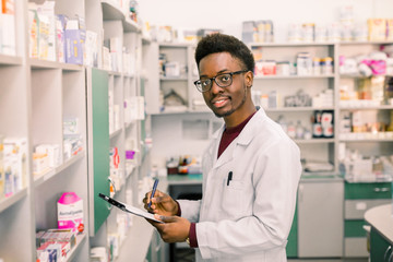 Un pharmacien ou un chimiste afro-américain souriant écrit sur le presse-papiers en se tenant debout à l& 39 intérieur de la pharmacie