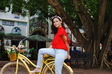 Outdoor portrait of young beautiful girl 19 to 25 years old posing in street. Brunette. riding a retro styled bicycle. Wearing red blouse With a penetrating camera look. 