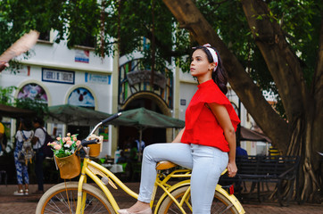 Outdoor portrait of young beautiful girl 19 to 25 years old posing in street. Brunette. riding a retro styled bicycle. Wearing red blouse With a penetrating camera look. 