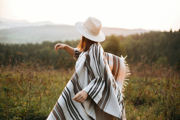 Stylish boho girl walking in sunny light at atmospheric sunset in meadow. Happy hipster woman in poncho and hat enjoying traveling in evening mountains. Space for text - obrazy, fototapety, plakaty