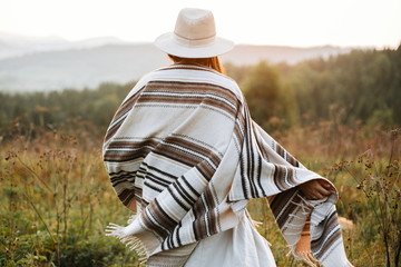 Stylish boho girl walking in sunny light at atmospheric sunset in meadow. Happy hipster woman in...