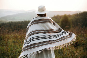Happy hipster woman in poncho and hat enjoying traveling in evening mountains. Stylish boho girl walking in sunny light at atmospheric sunset in meadow. Space for text