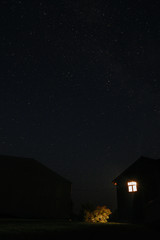 Beautiful view at night sky with stars and wooden cabin with light inside at trees on hill. Starry night in summer mountains. Wanderlust and travel concept