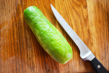 One big green fresh cucumber with a metal knife on a brown wooden kitchen board.