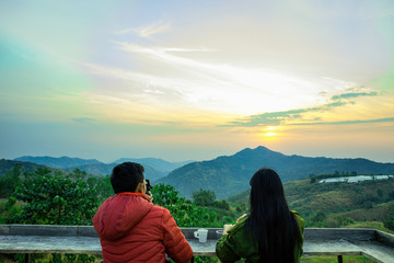 Young couple are looking beautiful sunrise moment on the mountain, Beautiful and romantic scene of couple are relax with landscape mountain and flare light in the morning