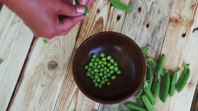 closeup of woman hand depod green peas in brown dish, on wooden table.