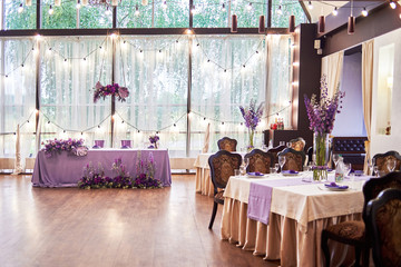Wedding presidium in restaurant, copy space. Banquet table for newlyweds with flowers, monstera leaves, violet cloth and bulbs. Lush floral arrangement. Luxury wedding decorations