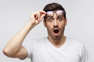 Young man in white t-shirt shouting oh my god with open mouth, surprised by low price and sales, holding glasses, isolated on gray background - obrazy, fototapety, plakaty