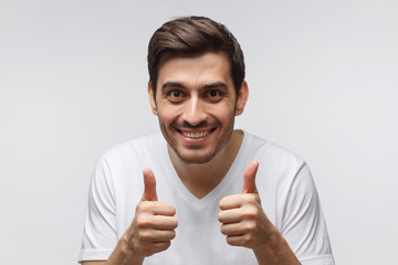 Excited young man in white tshirt making a thumbs up gesture of approval and success with smile,...