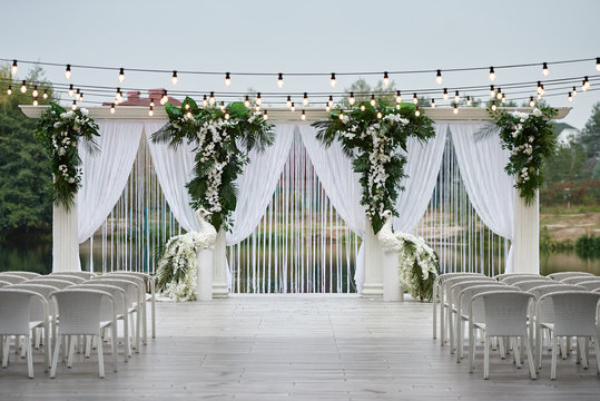 Place for wedding ceremony with wedding arch decorated with palm leaves, orchid flowers and floral peacocks, bulbs garland and white chairs outdoors, copy space