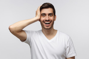 Handsome surprised man looking at the camera, astonished with big sale prices, holding hand on head, standing against gray background. Human face expression