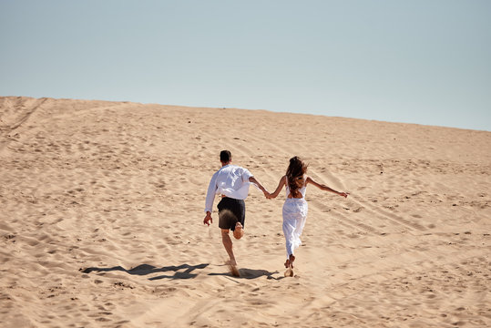 Happy loving couple holding hands and running in desert, copy space. Young lovers together on sands in summer - Powered by Adobe