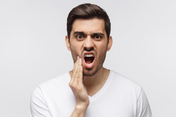Toothache concept. Close-up of young man isolated on gray background touching his face with expression of horrible suffer from health problem and aching tooth