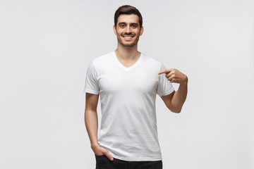 Young man pointing with index finger at blank  t-shirt with empty space for your advertising text or image, standing isolated on gray background - obrazy, fototapety, plakaty