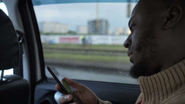 Portrait of black man is browsing smartphone. He is riding a car in rainy day in city. Afro american bearded guy is sitting in the back seat, side view.