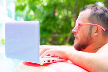 serious Fat funny man in pink inflatable circle in pink glasses works on a laptop in a swimming pool