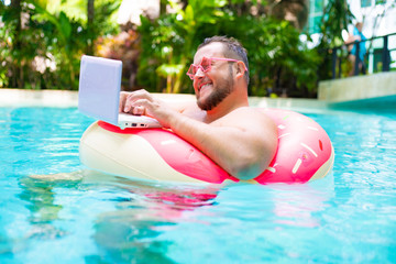 laughing Fat funny man in pink inflatable circle in pink glasses works on a laptop in a swimming pool