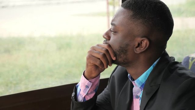 Afro american pensive tired young businessman is sitting in cafe. Portrait of young black man looks at window turns his head and looks at camera. Bearded young guy wears shirt and suit jacket.
