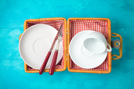 White Dishes And Tableware In Picnic Basket On Blue Wooden Background. Top View From Above