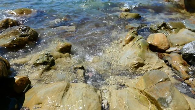 Sea surf on big rocks. Water waves on beach with multicolored polished stones, closeup view. Sunny summer day. Natural background.