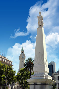 Vertical view of The Piramide de Mayo in Buenos Aires, Argentina, surrounded by old buildings and green palm trees, against a blue summer sky.