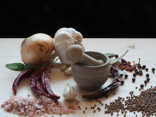 Mortar and pestle, surrounded by herbs, spices, onion, garlic and chili. Light wooden table and black top background.