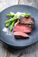 Barbecue dry aged wagyu sirloin beef steak sliced green asparagus and parmesan cheese as closeup on a gray cast iron design plate
